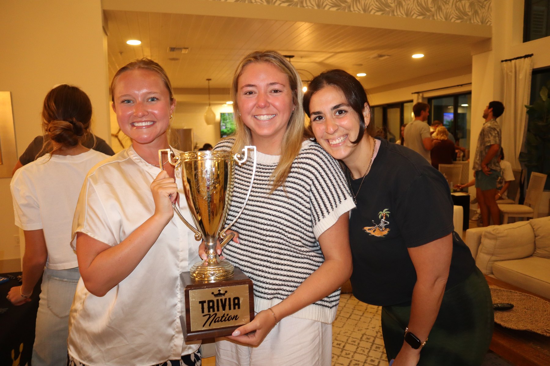 Three friends smiling and holding the Trivia Nation trophy at a private house party