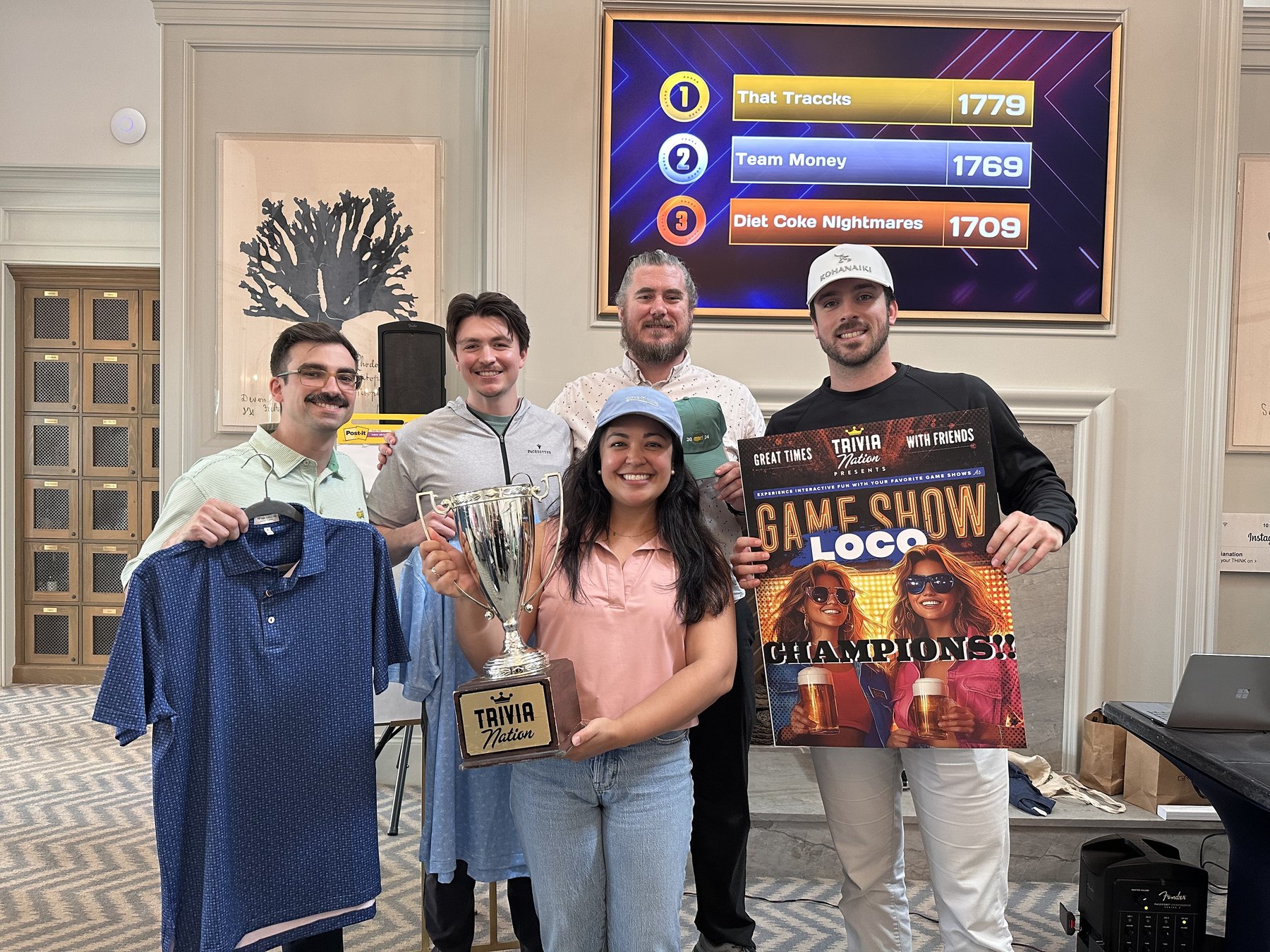 Winning team holding the Trivia Nation trophy and a Game Show Loco champions poster in front of the live scoreboard