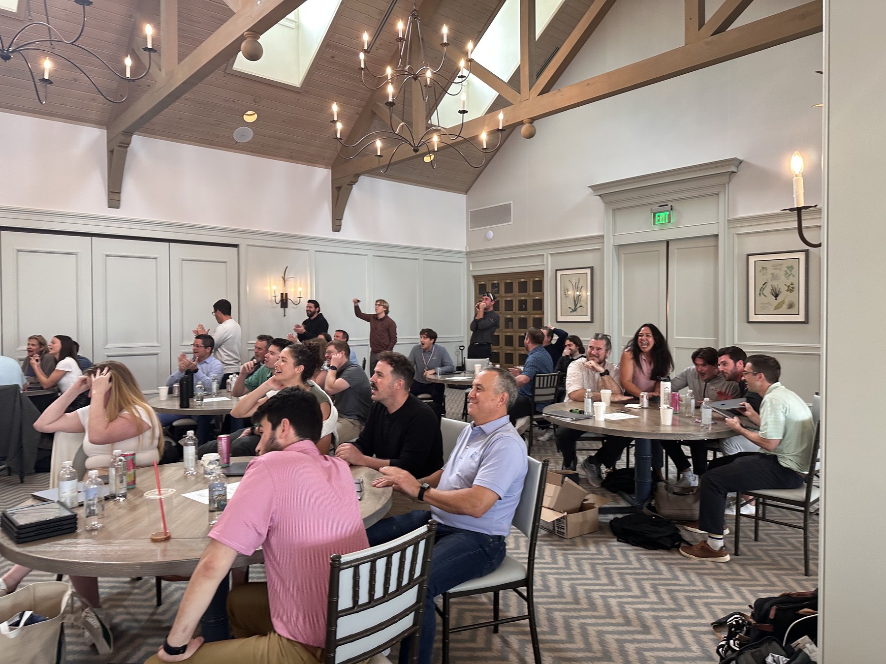 Club members seated at round tables in a vaulted clubhouse room laughing during a live-hosted game